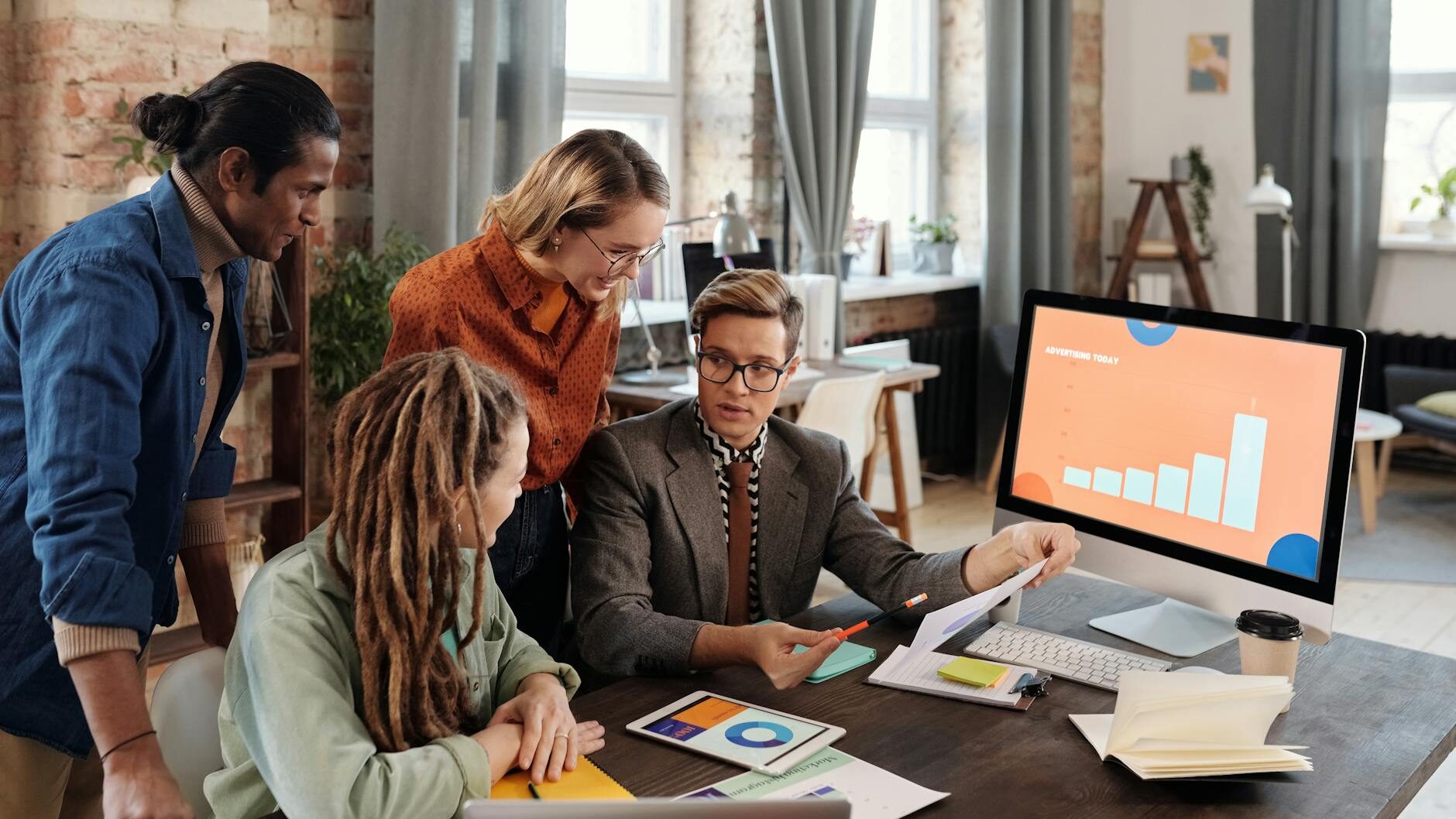 ビジネスミーティング中の4人の人々がデスクの周りに集まっている。1人がコンピュータの画面を指し示している。